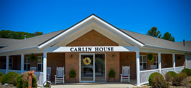 Front exterior view of Carlin House Assisted Living facility with a brick facade, white trim, a covered porch with white rocking chairs, potted plants, and a clear blue sky above.