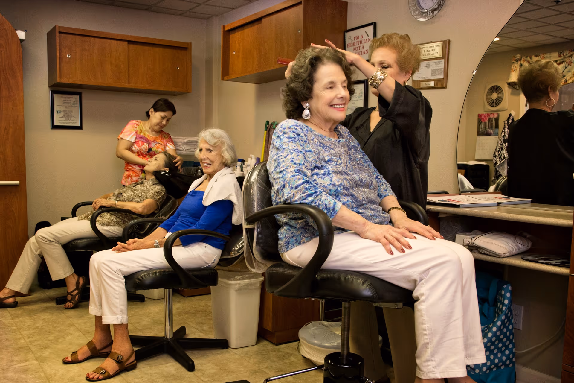 Two elderly women seated in salon chairs receiving hair styling services from two hairdressers in a cozy salon room with wooden cabinets and a large mirror.