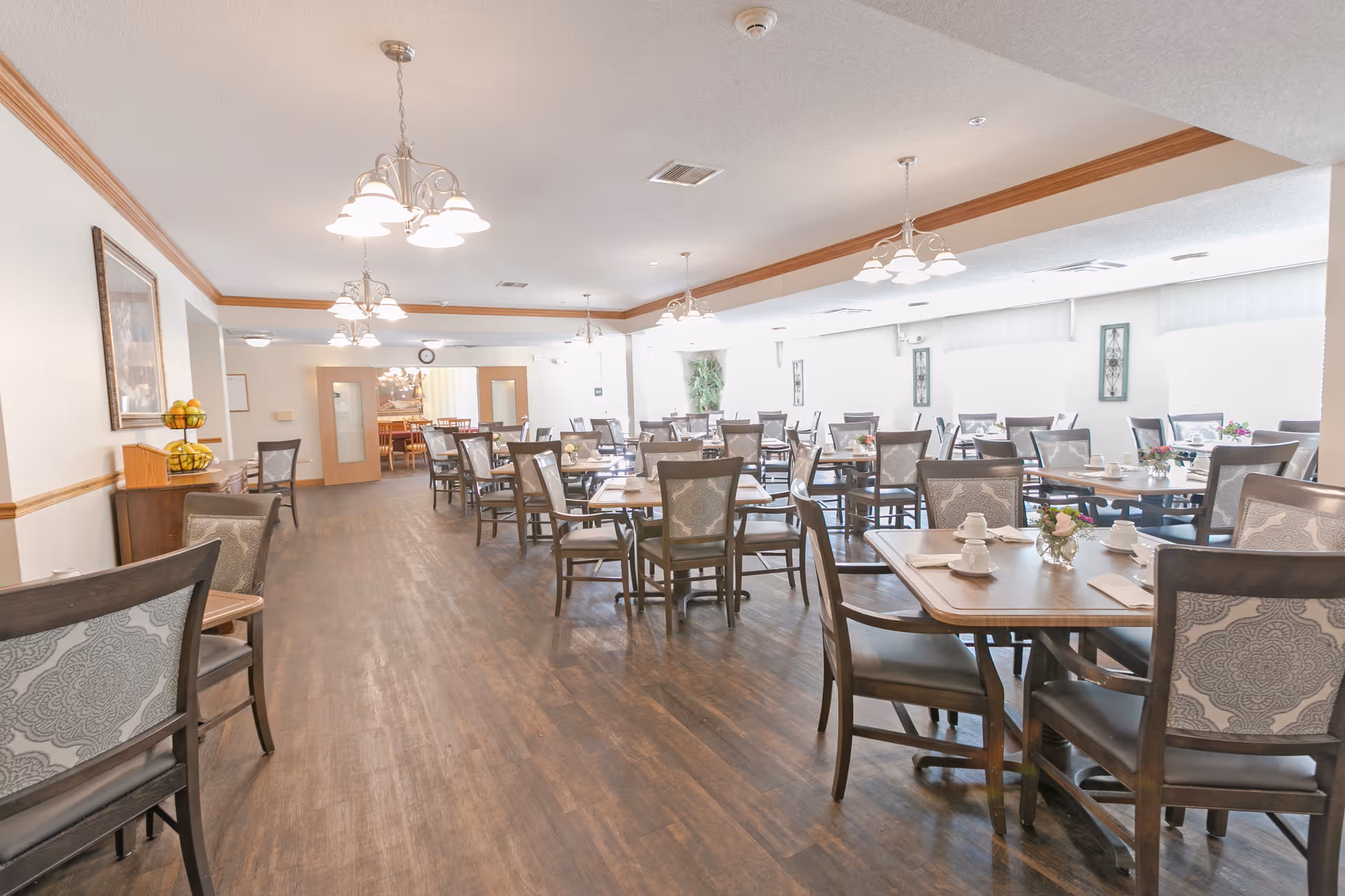 A spacious dining room with multiple wooden tables and chairs arranged neatly. Each table is set with white cups, saucers, and small flower arrangements. The room has wooden flooring, white walls with wooden trim, and several hanging light fixtures. There are framed artworks on the walls and large windows allowing natural light to brighten the space.