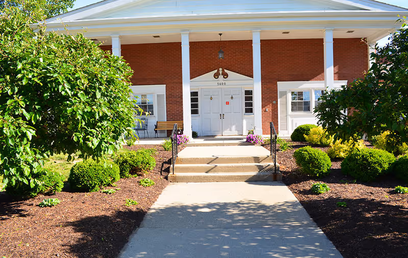 Front entrance of a brick building with white columns and a white double door, surrounded by landscaped bushes and trees with a concrete pathway leading up to the steps.