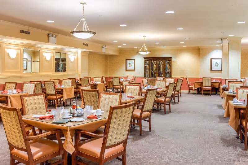A spacious dining room with multiple wooden tables and chairs arranged neatly. Each table is set with white cups, plates, glasses, and red napkins. The room has beige walls with framed artwork and a wooden cabinet against the far wall. Ceiling lights and wall sconces provide warm lighting.