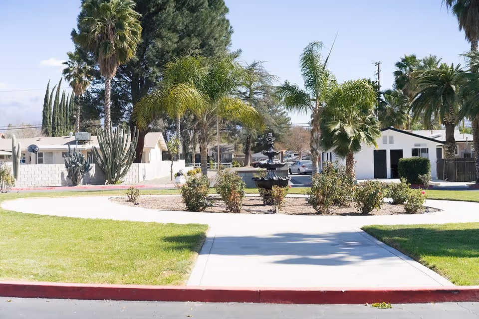 Outdoor view of a senior living facility with a circular garden area featuring a multi-tiered black fountain surrounded by palm trees and shrubs. There is a paved walkway leading to the garden, green grass on either side, and residential buildings in the background under a clear sky.