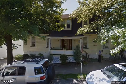 Front exterior view of a two-story residential building partially obscured by trees, with a porch and two vehicles parked on the street in front.