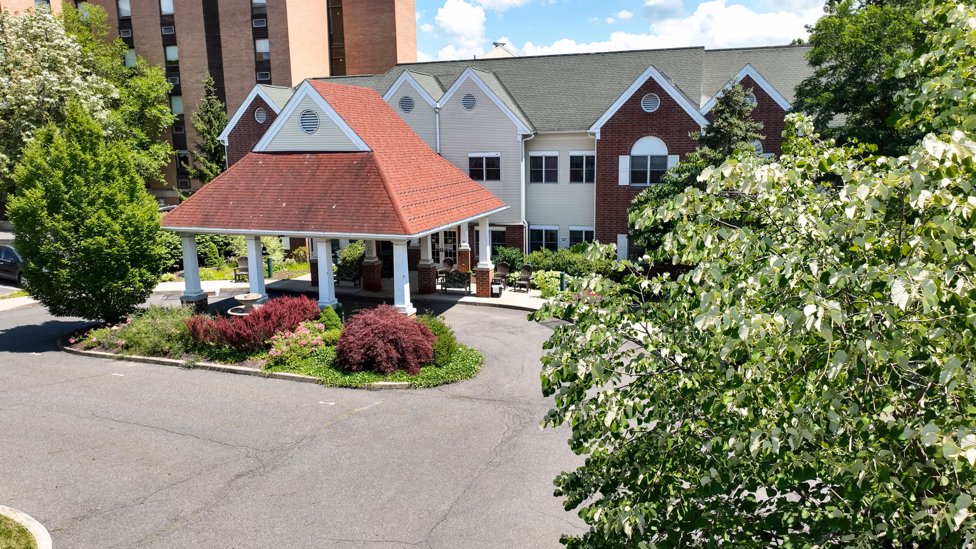Exterior view of Allied Services Meade Street Residence showing a covered entrance with a red roof, surrounded by greenery and bushes, with a two-story building in the background under a partly cloudy sky.