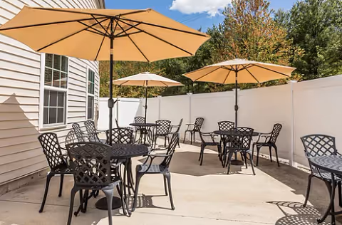 Outdoor patio area with several black metal tables and chairs, each table shaded by a beige umbrella. The patio is enclosed by a white fence and adjacent to a beige building with windows. Trees and blue sky are visible in the background.