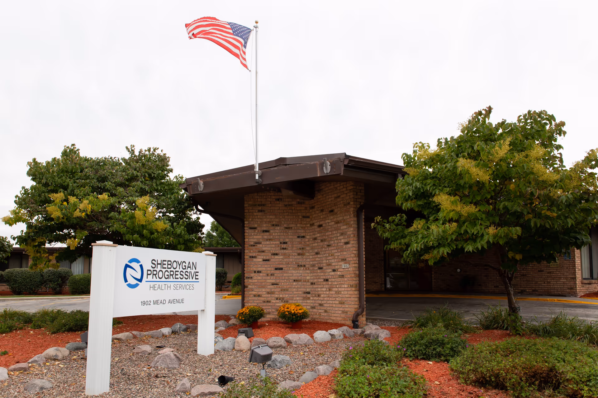 Exterior view of Sheboygan Progressive Health Services building with a brick facade, an American flag flying on a pole, landscaped garden with bushes, trees, and rocks, and a white sign displaying the facility name and address.