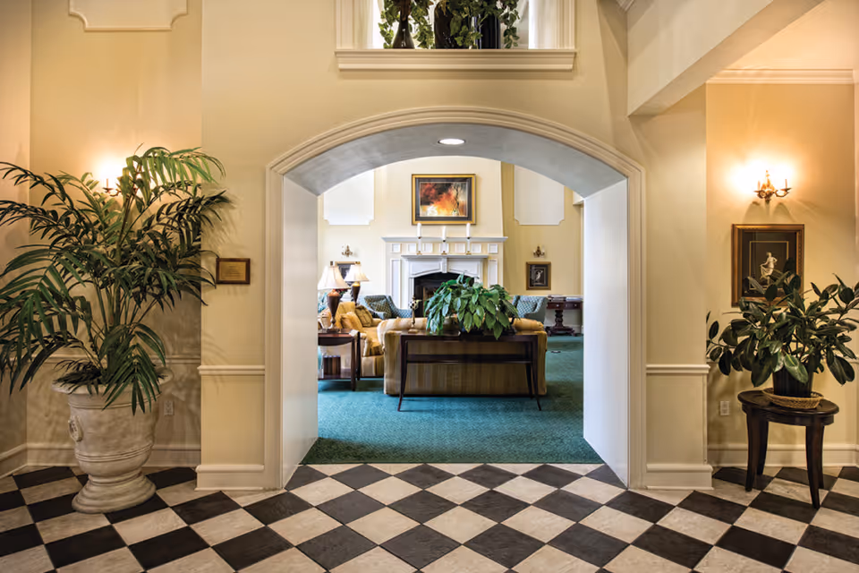 View through an arched doorway into a cozy living room area with a fireplace, sofas, armchairs, lamps, and framed artwork on the walls. The foreground features a black and white checkered floor with large potted plants on either side of the archway.