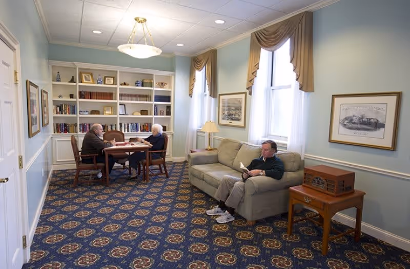 A cozy common room with patterned carpet, light blue walls, and two large windows with beige curtains. Two elderly people are sitting at a wooden table with books, while another person is seated on a beige couch reading. The room has a built-in white bookshelf filled with books and decorative items, framed artwork on the walls, a table lamp, and a wooden side table with a vintage-style radio.
