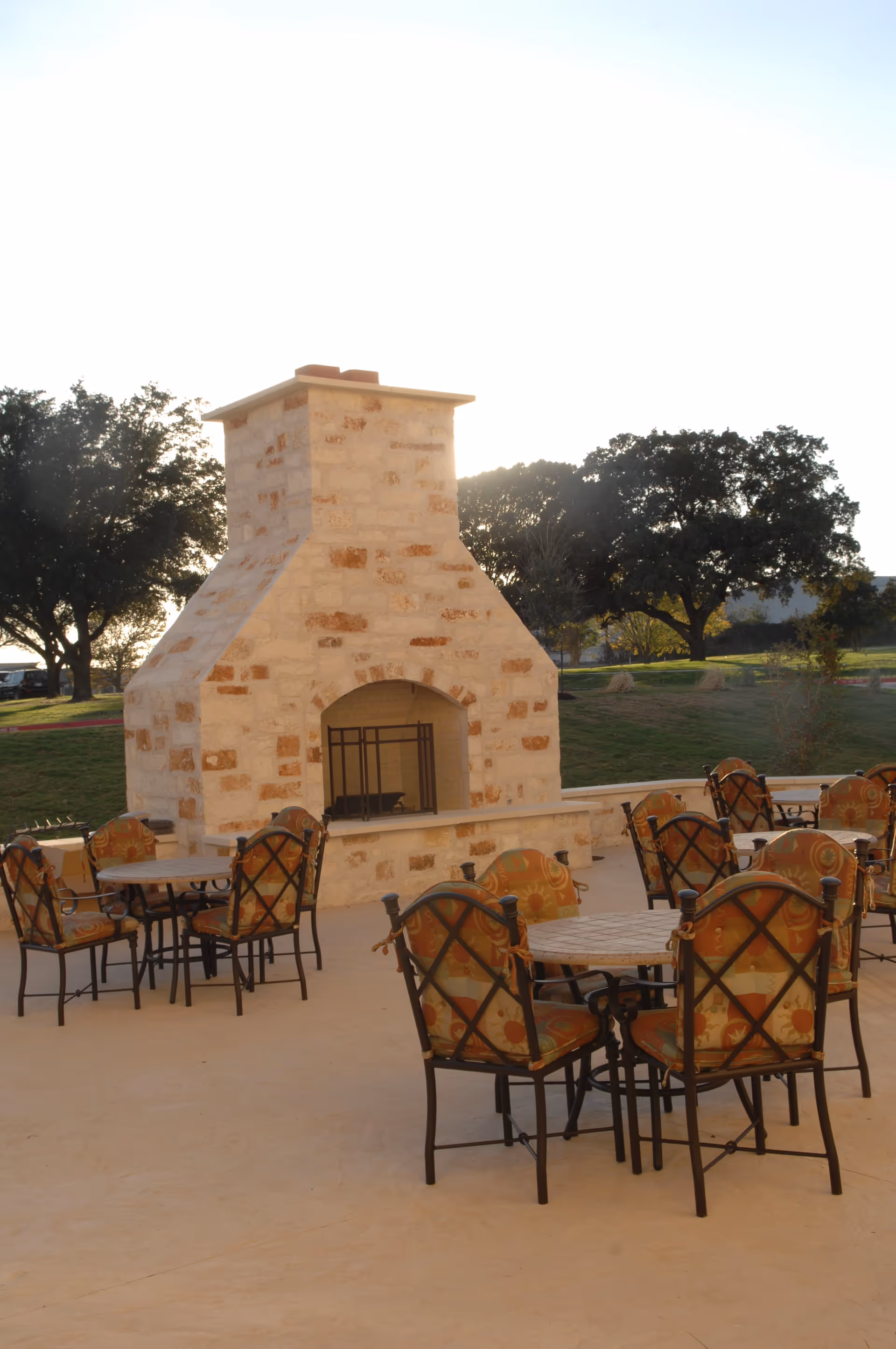 Outdoor patio area with multiple round tables and cushioned chairs arranged around a large stone fireplace. Trees and grassy areas are visible in the background under a bright sky.