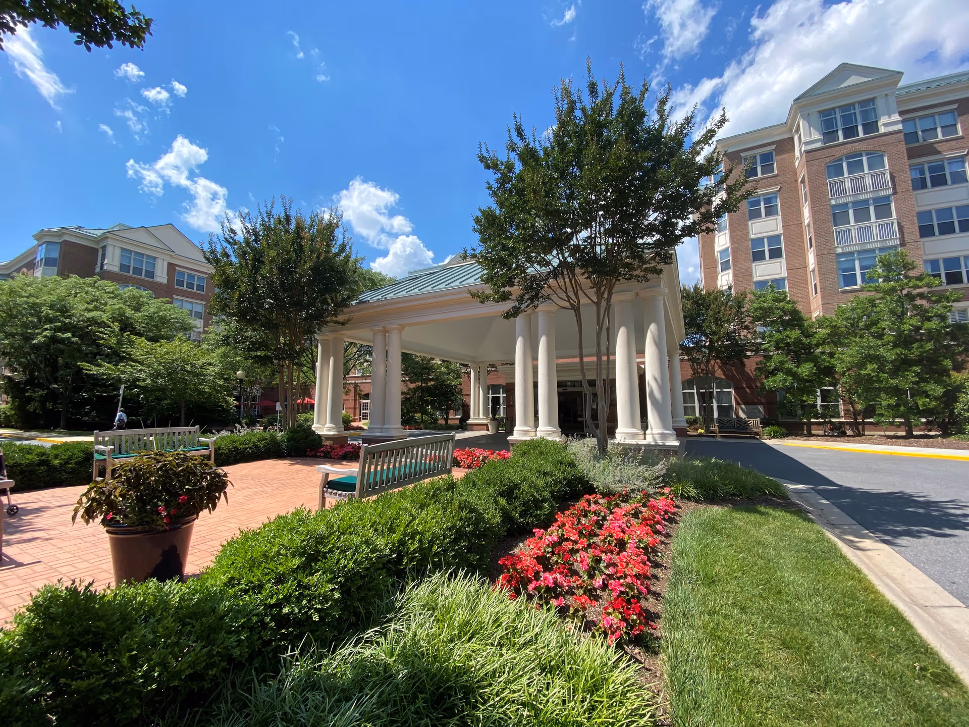 Outdoor view of Maplewood Park Place featuring a covered entrance with white columns, surrounded by well-maintained landscaping including green bushes, red flowers, and trees. The building is a multi-story brick structure under a blue sky with some clouds.
