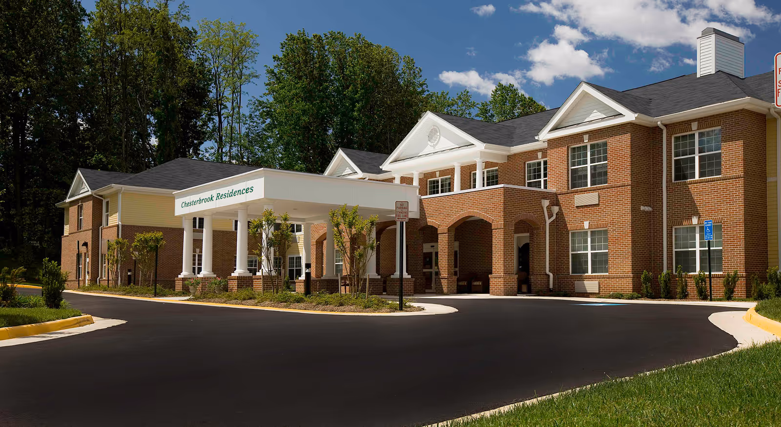 Exterior view of Chesterbrook Residences, a two-story brick building with white columns and a covered entrance. The building is surrounded by trees and landscaping under a partly cloudy blue sky.