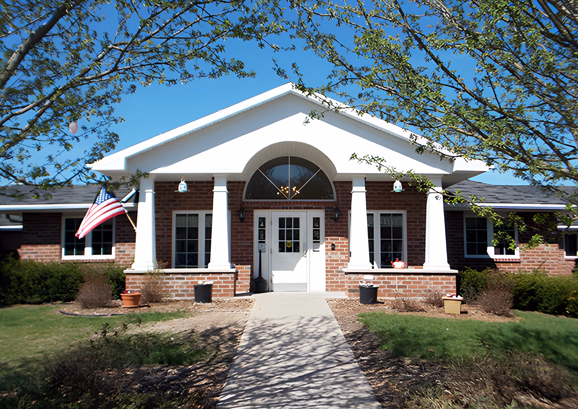 Front exterior view of a single-story brick building with white columns and a white door entrance. There is an American flag mounted on the left side and some small potted plants near the entrance. Trees with green leaves frame the building under a clear blue sky.