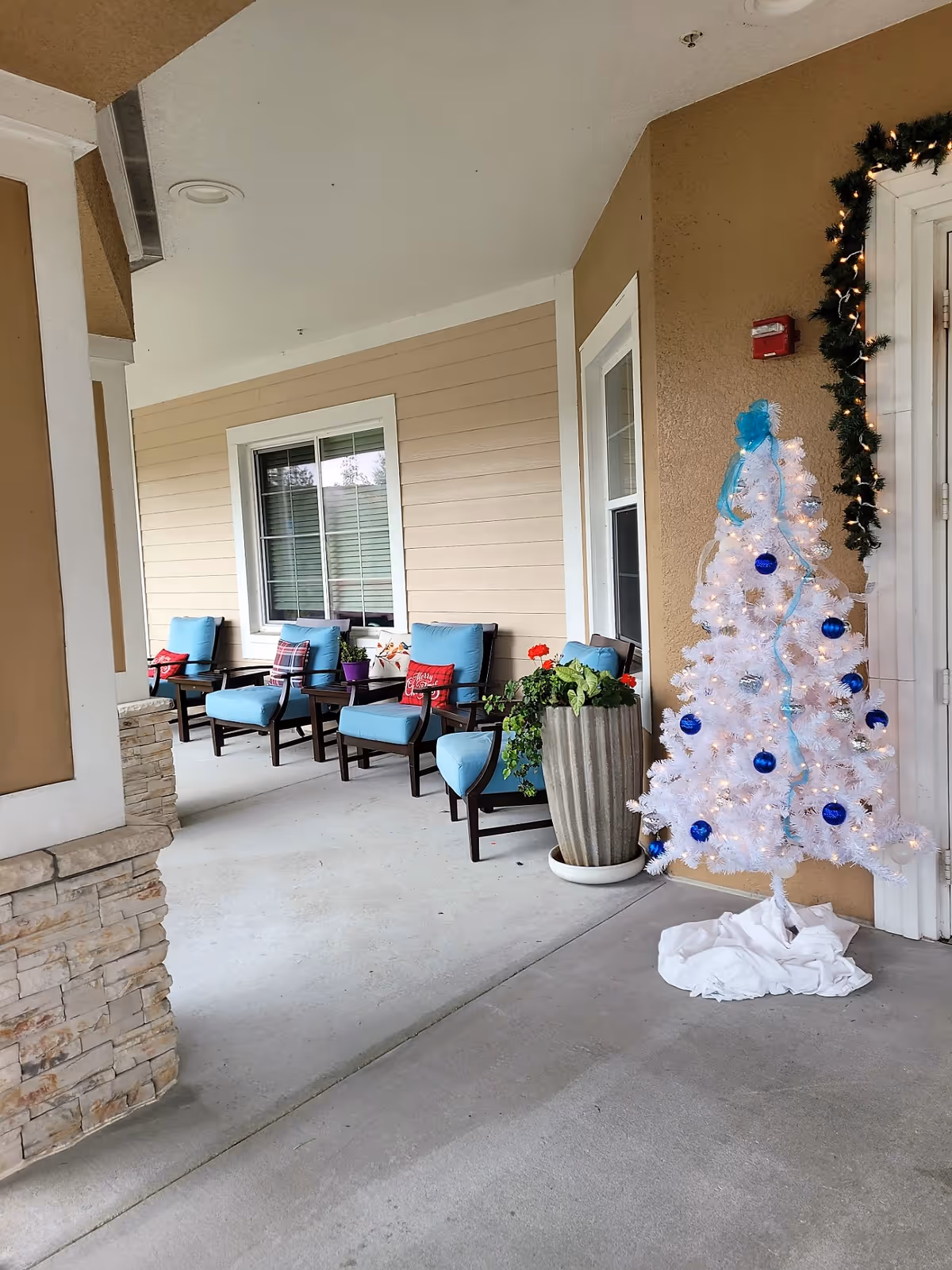 Covered outdoor seating area with four cushioned chairs in blue, small tables, potted plants, and a white Christmas tree decorated with blue ornaments and lights near a door. The area has beige walls and a stone pillar.