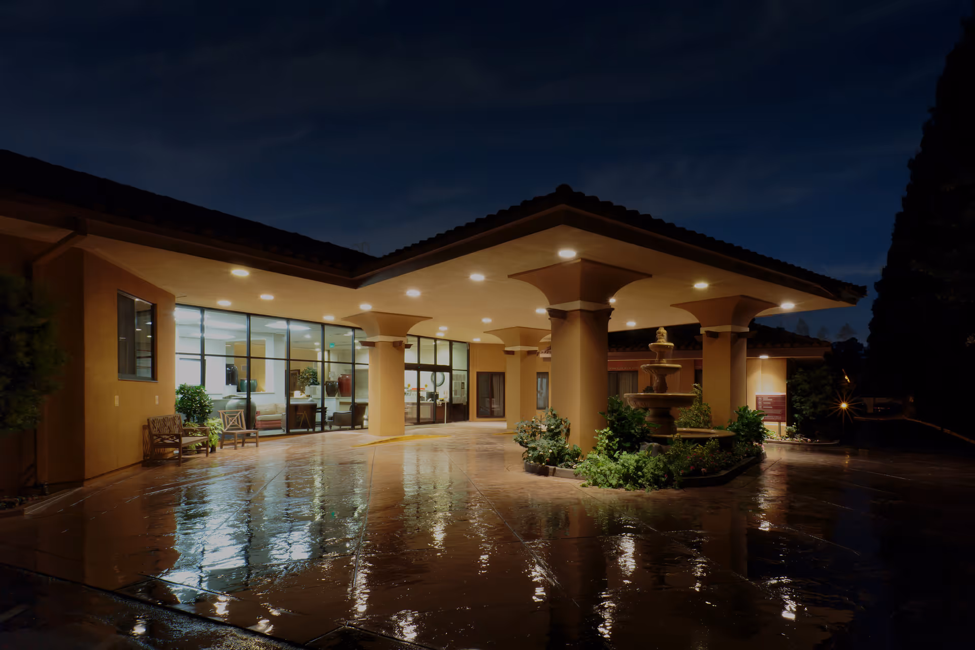 Night view of the entrance to Vasona Creek Healthcare Center, featuring a covered driveway with columns, a central fountain surrounded by greenery, and large glass windows showing the interior seating area.