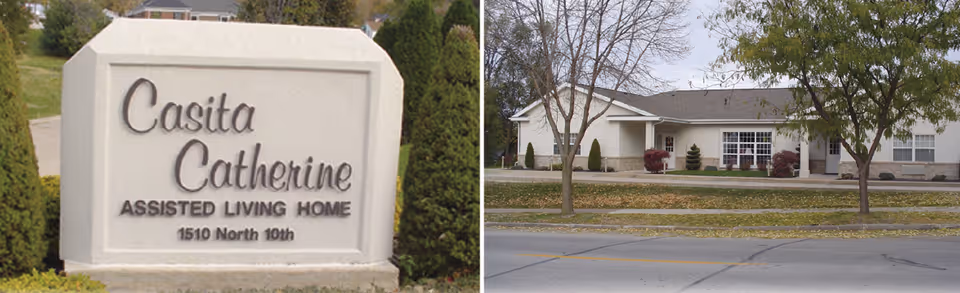 A large stone sign reading 'Casita Catherine Assisted Living Home 1510 North 10th' is positioned on a grassy area with bushes around it. Across the street, there is a single-story building with white exterior walls, several windows, and a few trees and shrubs in front.