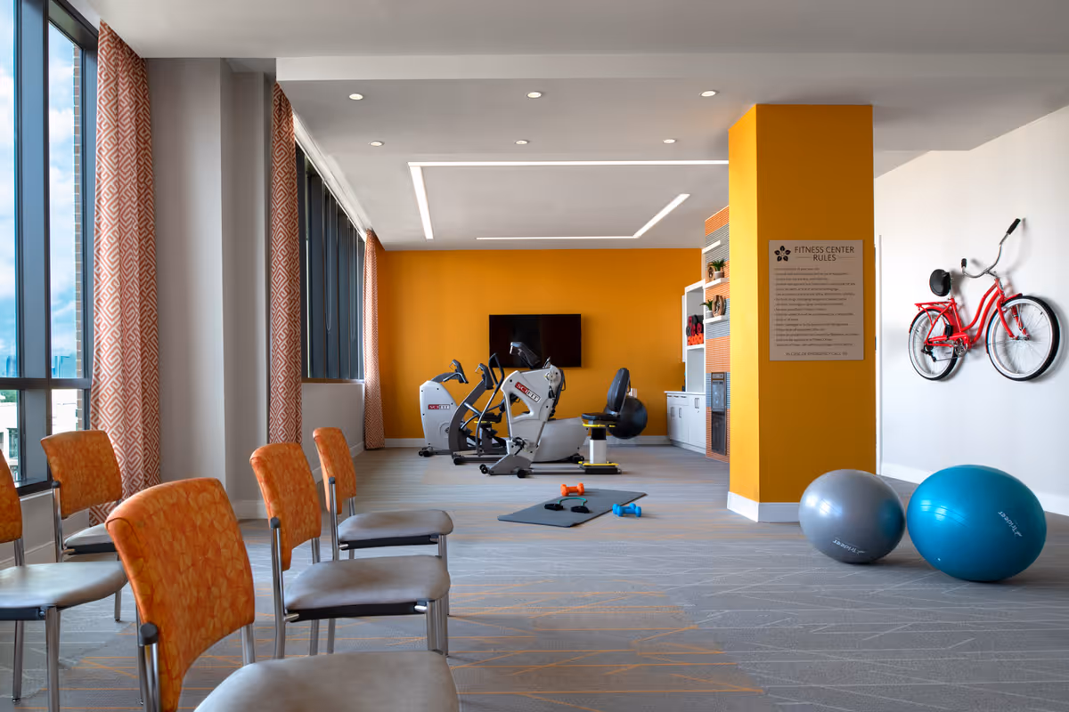 A bright fitness center with large windows and orange patterned curtains. The room features exercise equipment including stationary bikes, exercise balls, dumbbells on a mat, and a red bicycle mounted on the wall. There are orange and gray chairs arranged in the foreground, and a yellow accent wall with a mounted TV and a sign listing fitness center rules.