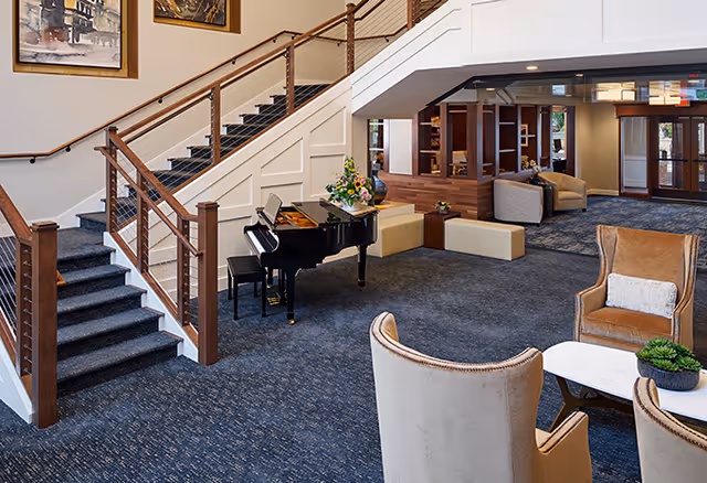 Interior view of a senior living facility common area featuring a grand piano near a staircase with wooden railings, comfortable seating including armchairs and sofas, a small table with a plant, and decorative artwork on the walls.
