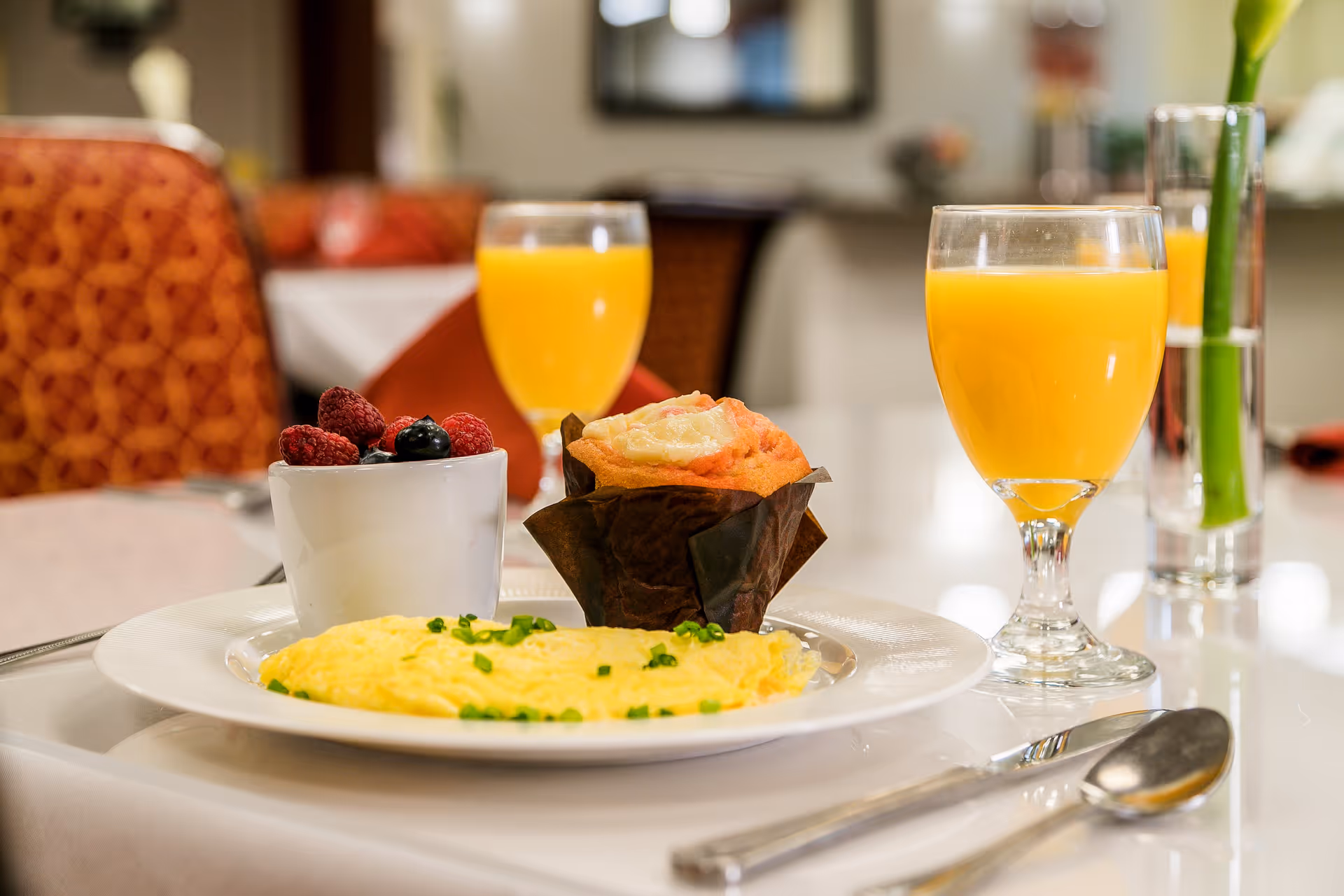 A breakfast plate with a folded omelette garnished with chopped chives, a muffin in a brown wrapper, and a small cup of mixed berries. Two glasses of orange juice and a vase with a single yellow flower are also on the table. The setting is in a dining area with patterned orange chairs and a softly blurred background.
