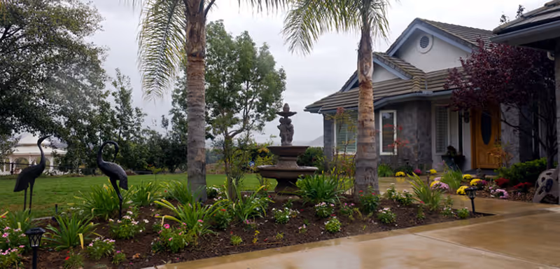 A landscaped garden area in front of a residential building with a stone fountain, palm trees, various plants, flowers, and two decorative bird statues. The building has a tiled roof and a yellow front door.