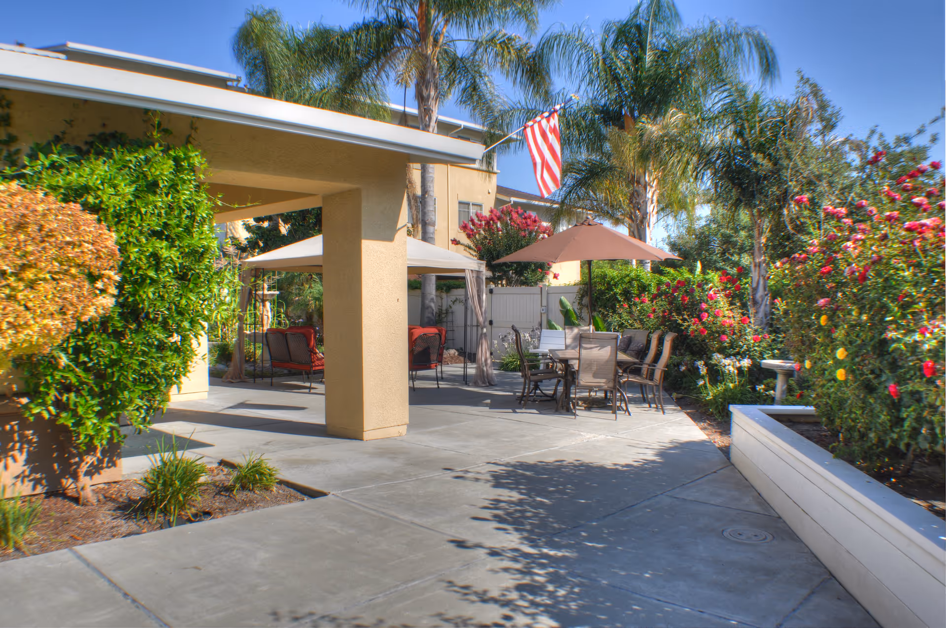 Outdoor patio area at Carlton Senior Living San Jose with seating arrangements including chairs and tables under umbrellas, surrounded by lush greenery, palm trees, and flowering bushes, with an American flag displayed on the building.