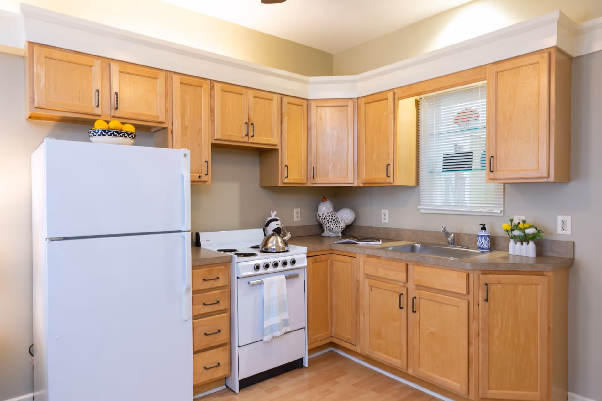 A clean kitchen with light wooden cabinets, a white refrigerator, a white stove with a kettle on it, a countertop with a sink, a small window with blinds, and decorative items including a bowl of lemons, a ceramic rooster, a soap dispenser, and a small vase with yellow flowers.
