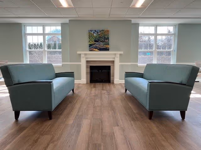 A bright room with two teal sofas facing each other on a wooden floor, a white fireplace centered on the far wall, and two large windows on either side of the fireplace letting in natural light. A colorful painting hangs above the fireplace.
