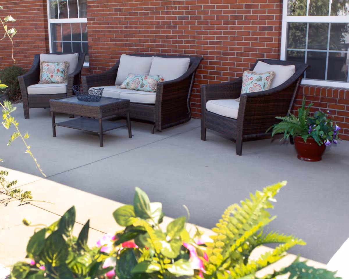 Outdoor patio area with a brick wall background featuring a wicker loveseat and two wicker armchairs with light-colored cushions and floral-patterned pillows. A wicker coffee table with a decorative basket sits in front of the seating. There are potted plants with green foliage and flowers in the foreground and to the side.