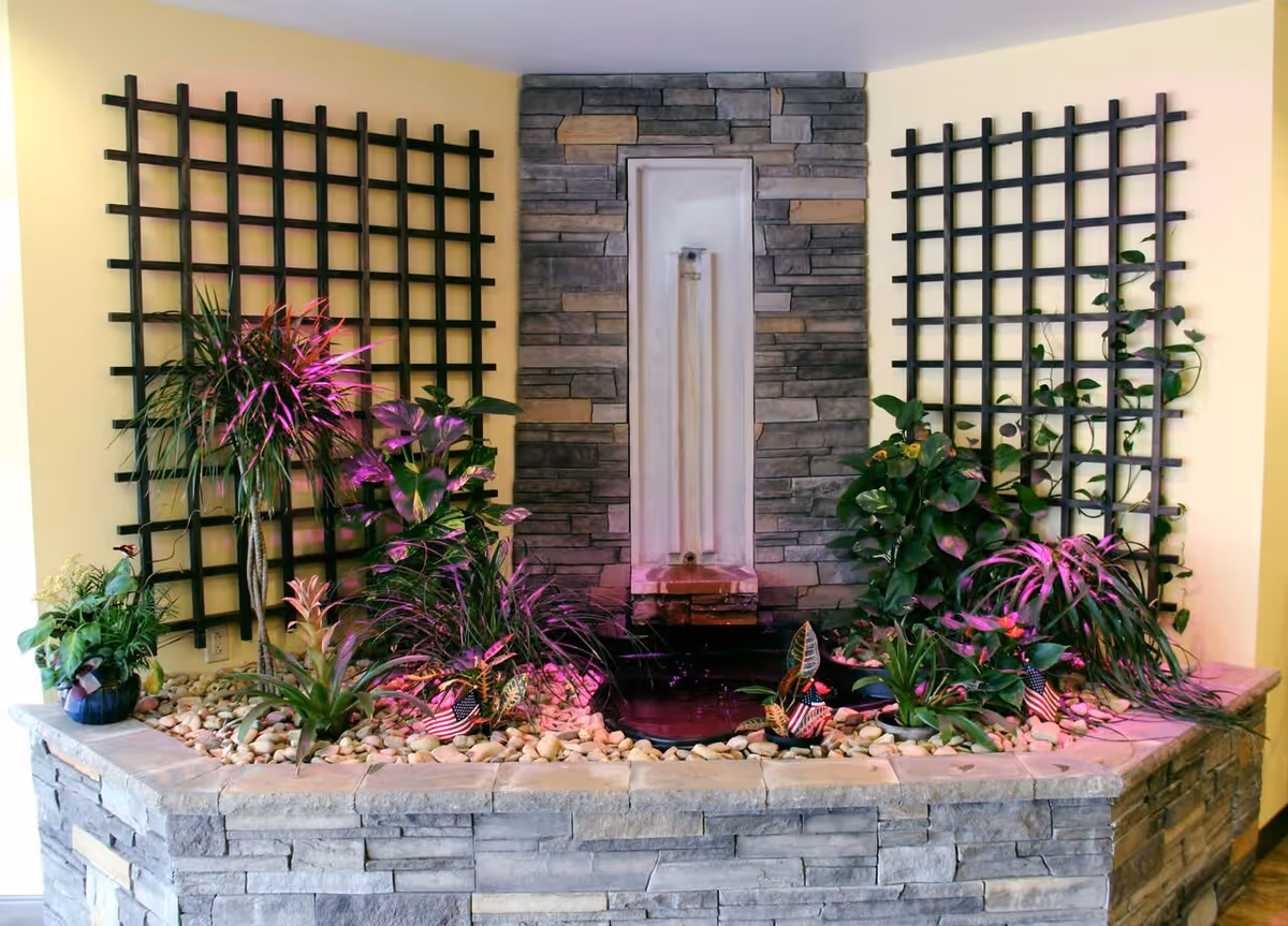 Indoor stone water fountain surrounded by various green plants and small American flags, with two black lattice panels mounted on the yellow walls behind it.