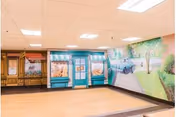 An indoor room with a wooden floor and a ceiling with recessed lighting. The walls feature a mural depicting a scenic outdoor setting with trees, a car, and a pathway. There is also a painted storefront facade with windows and awnings, and a wooden cabinet on the left side of the room.