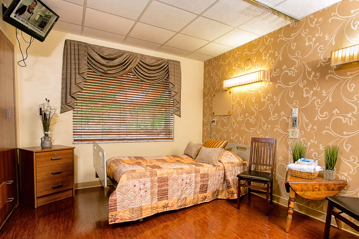 A cozy bedroom in a senior living facility with a single bed covered in a patchwork quilt, two pillows, a wooden chair, a small wooden table with a basket and plants, a wooden dresser with a vase of flowers, a window with blinds and decorative valance, and a wall-mounted TV in the corner.