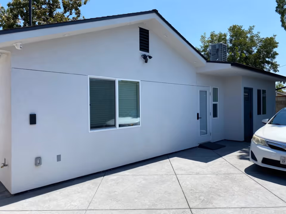 Exterior view of a single-story white building with a sloped roof, two windows, a door with a black handle, and a white car parked on a concrete driveway. Trees are visible in the background under a clear blue sky.