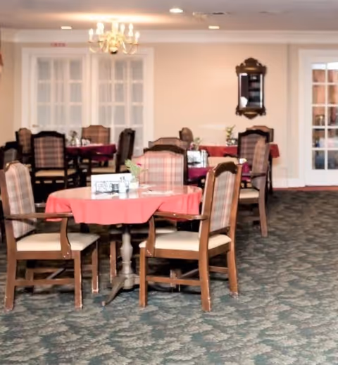 A dining room with several round tables covered with red tablecloths, each surrounded by wooden chairs with cushioned seats and plaid backs. The room has a patterned carpet, a chandelier hanging from the ceiling, and a mirror on the wall. French doors are visible in the background.