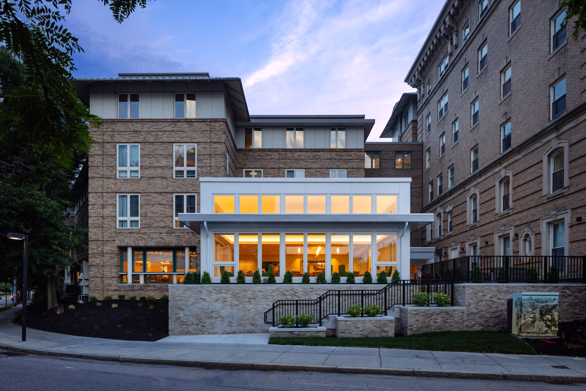 Exterior view of a multi-story brick senior living facility building at dusk with large windows illuminated from inside, showing a modern white extension with floor-to-ceiling windows and landscaped surroundings including stairs and greenery.