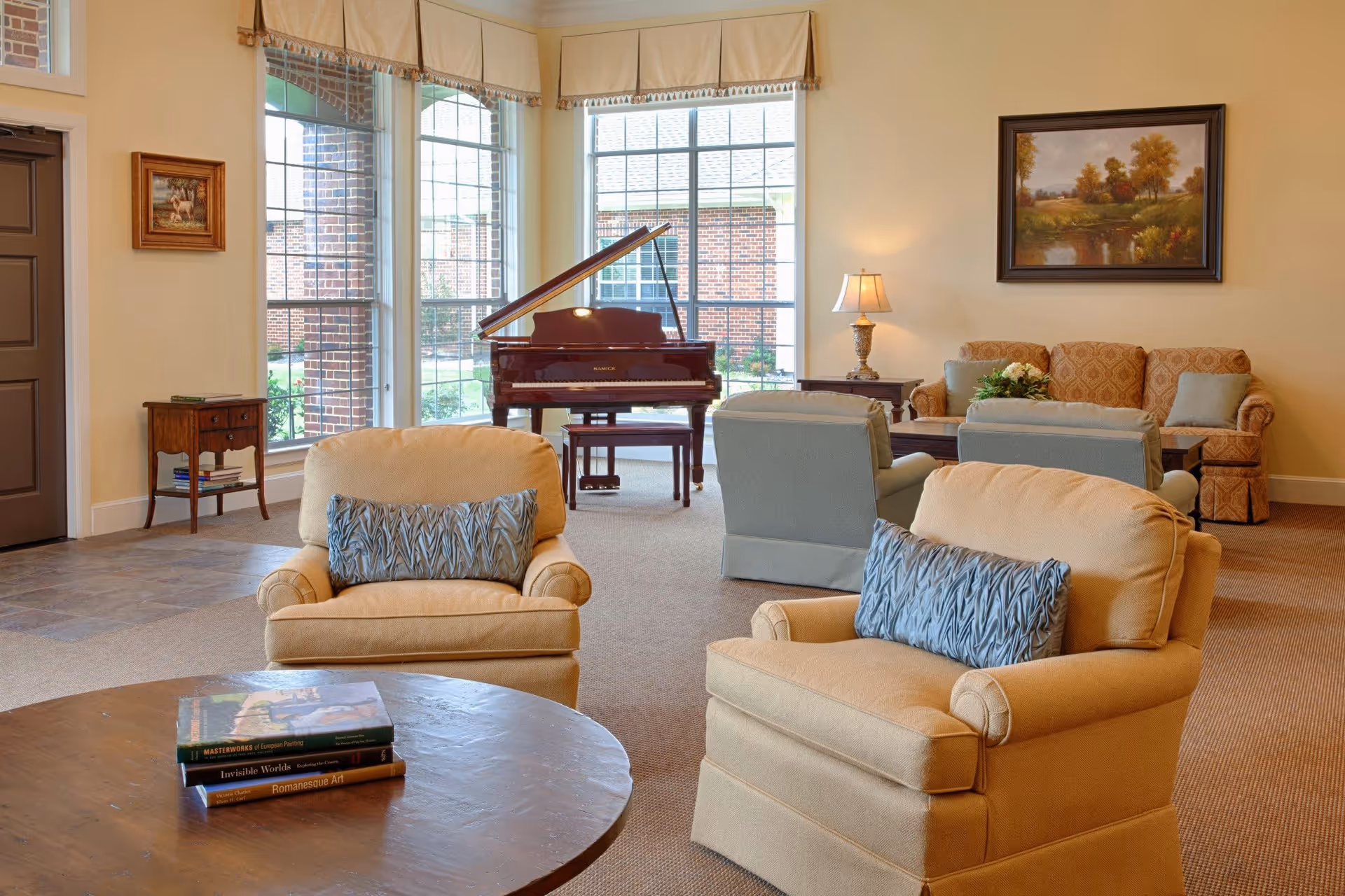 A cozy living room area with beige armchairs featuring blue patterned pillows, a round wooden coffee table with books, a grand piano near large windows, a sofa and two armchairs in the background, a side table with a lamp, and framed artwork on the walls.