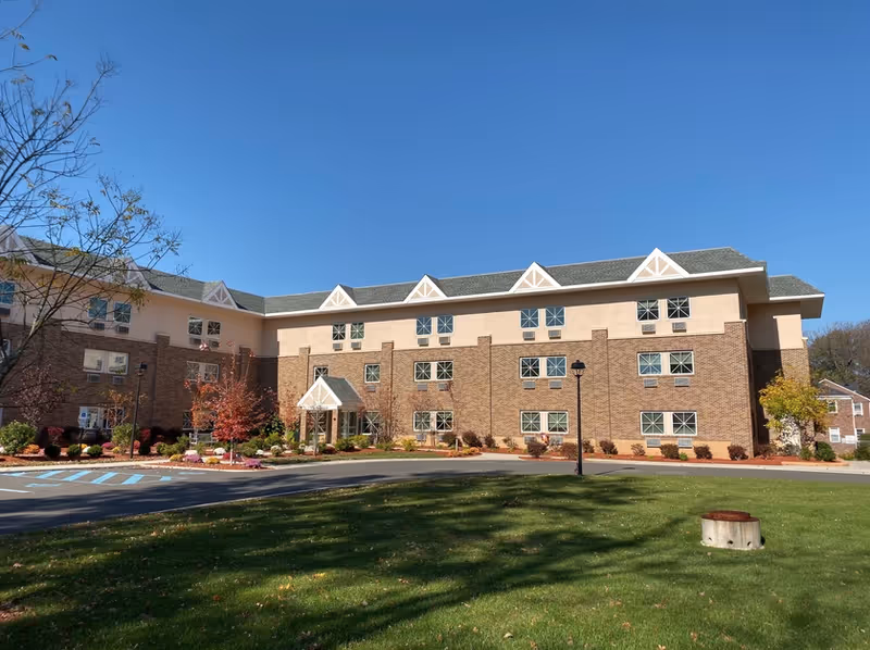Exterior view of a three-story senior living facility building with a brick and beige facade, multiple windows, a small entrance canopy, landscaped bushes and trees, a parking area with handicap parking spaces, and a green lawn in the foreground under a clear blue sky.