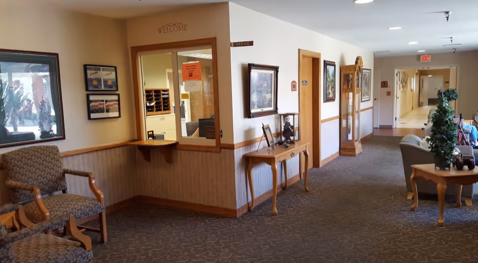Interior hallway of a senior living facility with patterned carpet, beige walls, and wooden trim. There are upholstered chairs on the left, a reception window labeled 'Office' with a 'Welcome' sign above it, framed pictures on the walls, a wooden table with decorative items, a grandfather clock, and a seating area with plants and more chairs further down the hallway.