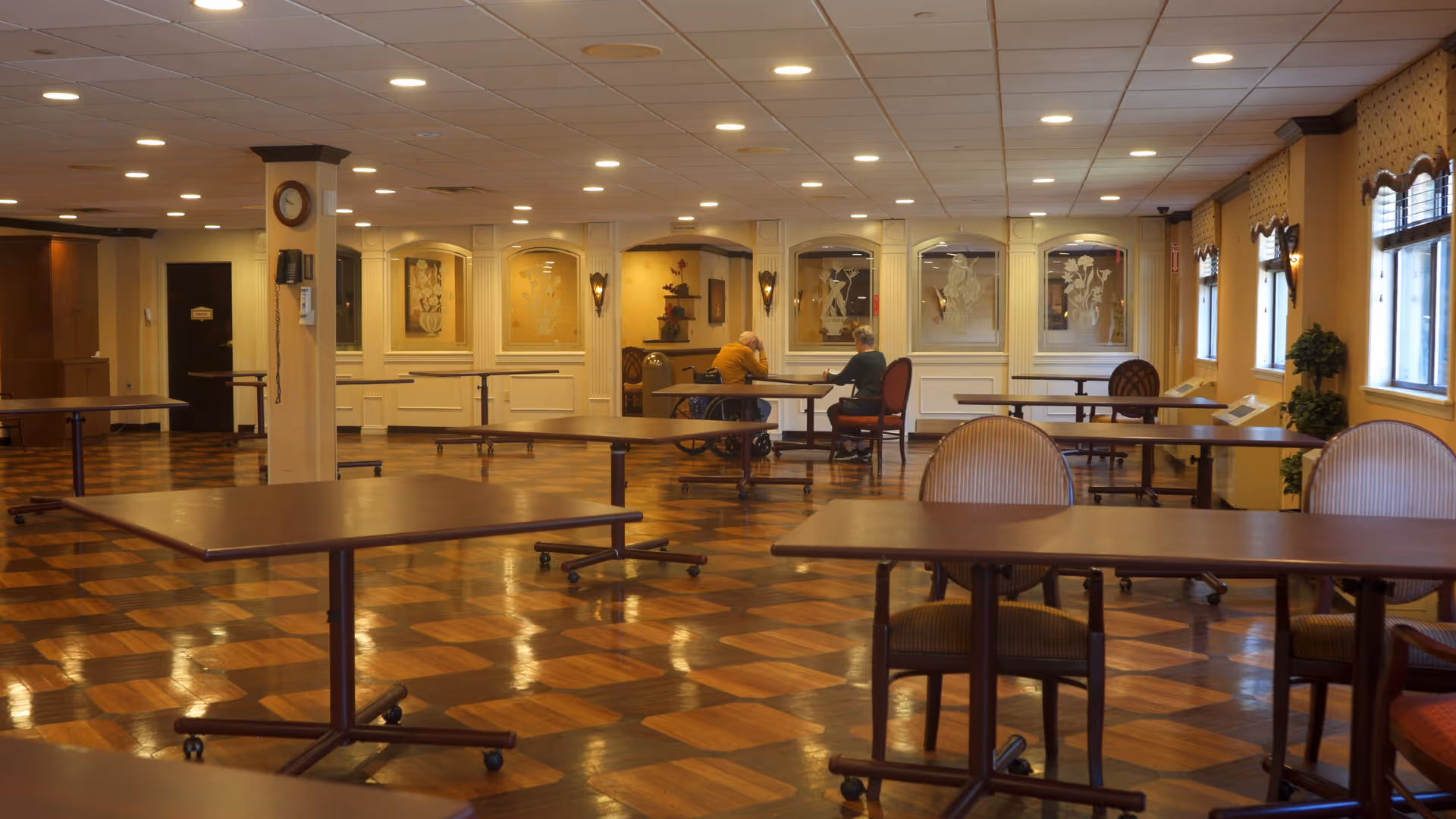 A spacious dining room with wooden tables and chairs arranged neatly on a polished checkered wood floor. Two elderly people are seated at a table near the back wall, which is decorated with framed artwork and sconces. Large windows on the right side let in natural light, and the ceiling has recessed lighting.