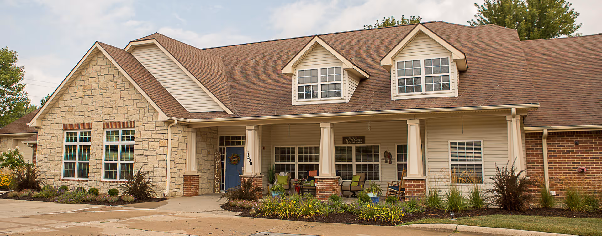 Front exterior view of a single-story senior living facility building with a combination of stone and brick facade, a brown shingled roof, and a covered porch with seating and welcome signs.