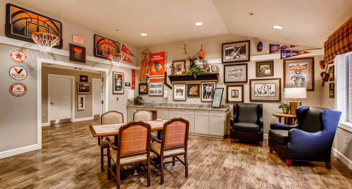 A cozy common area in Deer Ridge Memory Care decorated with sports memorabilia including basketball hoops, framed photos, and pennants. The room features a table with four chairs, two armchairs near a window with plaid curtains, and a countertop with cabinets underneath. The floor is wood, and the walls are painted beige.