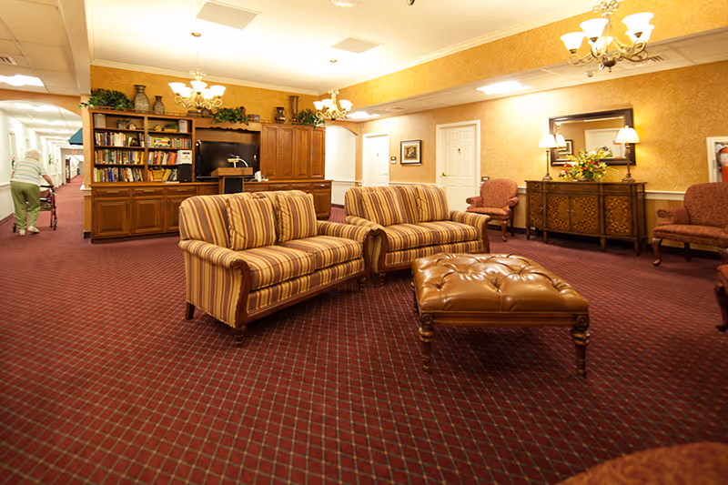 A cozy common area in a retirement community featuring two striped sofas, a brown leather tufted ottoman, and several upholstered chairs. The room has a red patterned carpet, warm yellow walls, a wooden bookshelf filled with books, and decorative plants. A person with a walker is seen in the hallway on the left side of the image.
