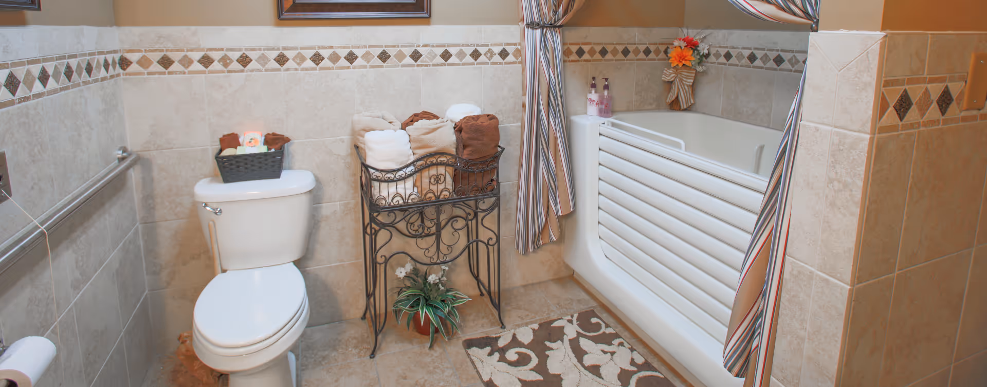 A bathroom with beige tiled walls and floor, featuring a white toilet with a basket of toiletries on top, a decorative metal stand holding rolled towels, a potted plant on the floor, and a white bathtub with a safety rail and striped shower curtains. There is a floral-patterned rug on the floor in front of the bathtub.