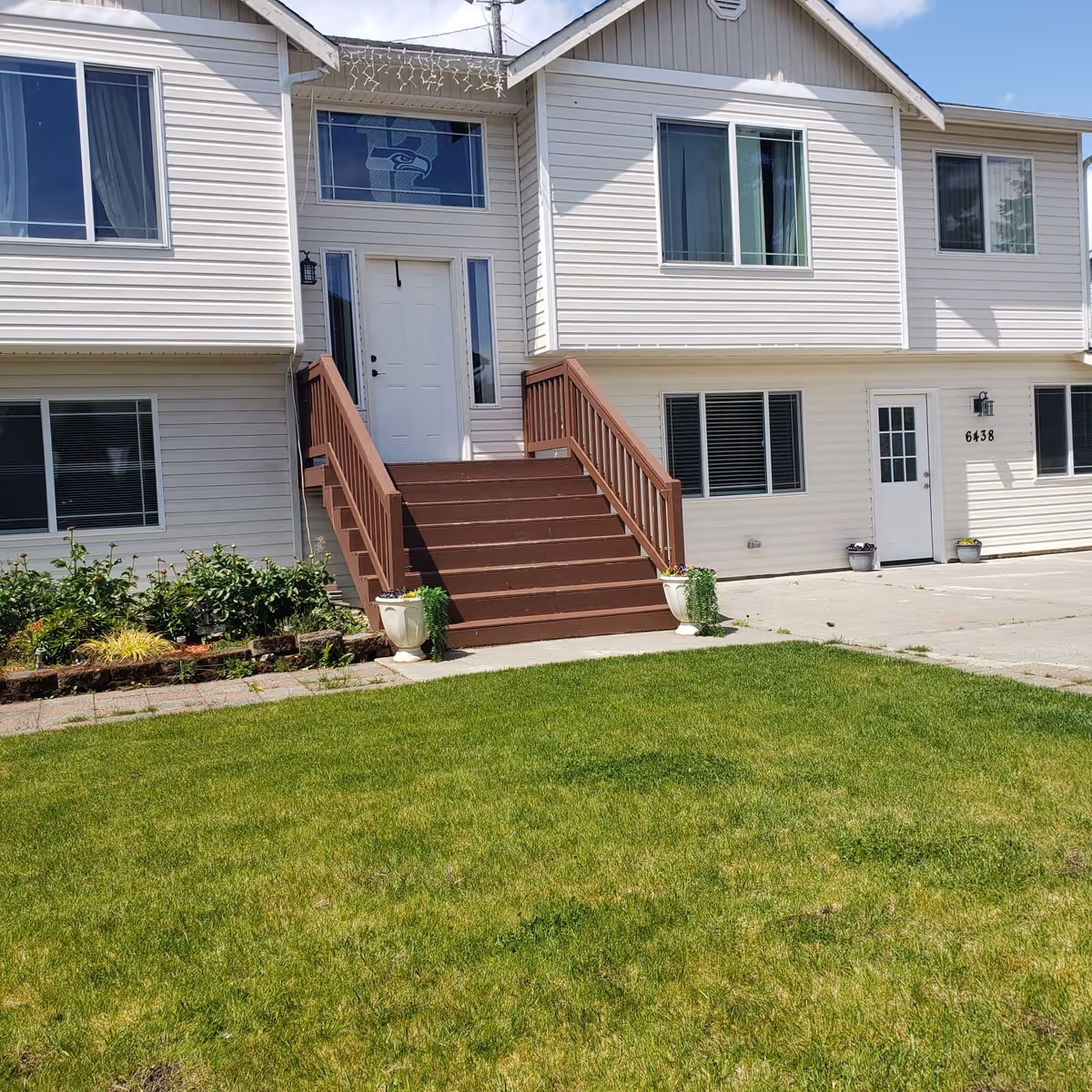 Front exterior view of a two-story white building with a brown wooden staircase leading to a white door. The building has several windows, a green lawn in front, and two white planters with plants on either side of the stairs. There is a decorative window above the door featuring a sports team logo.