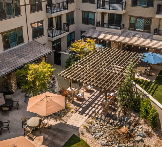 Outdoor courtyard area of a senior living facility featuring a wooden pergola with seating underneath, patio tables with umbrellas, landscaped garden beds with rocks and plants, and a multi-story building with balconies surrounding the courtyard.