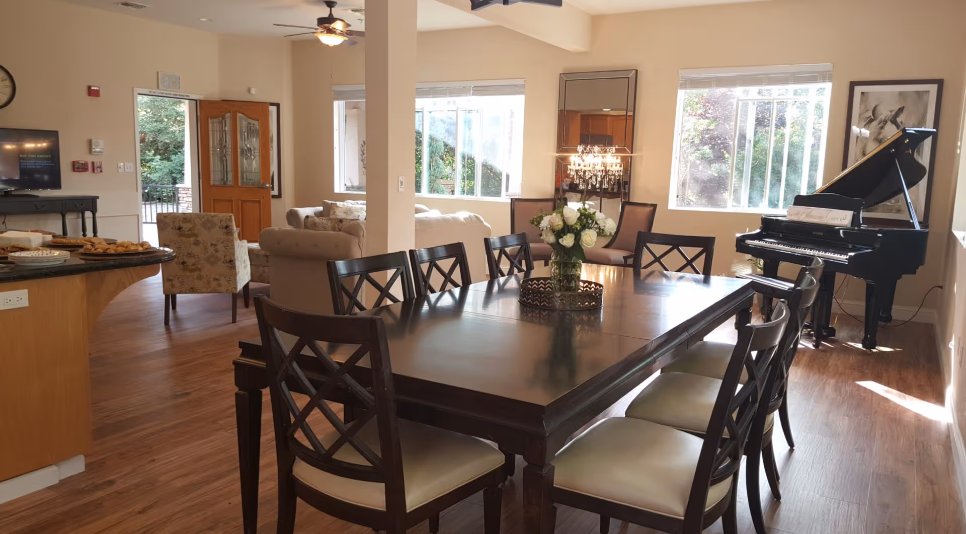 A bright and spacious common area in a senior living facility featuring a large wooden dining table with eight chairs, a vase of white flowers as a centerpiece, a black grand piano near the window, comfortable beige sofas and armchairs, a wooden door leading outside, and large windows letting in natural light.