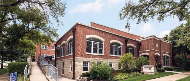 Exterior view of a two-story brick building with large windows, surrounded by trees and greenery. A paved walkway with a handrail and a handicap accessible sign leads to the entrance. The sky is partly cloudy.