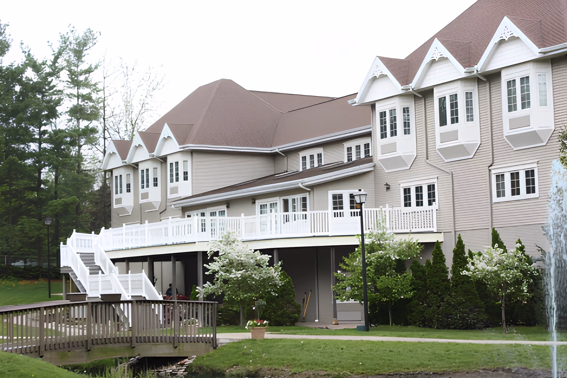 Exterior view of a senior living facility building with beige siding and a brown roof. The building features multiple white-framed windows and a white railing along an elevated walkway with stairs leading down to a garden area. There are green trees, bushes, and a small wooden bridge over a water feature in the foreground.