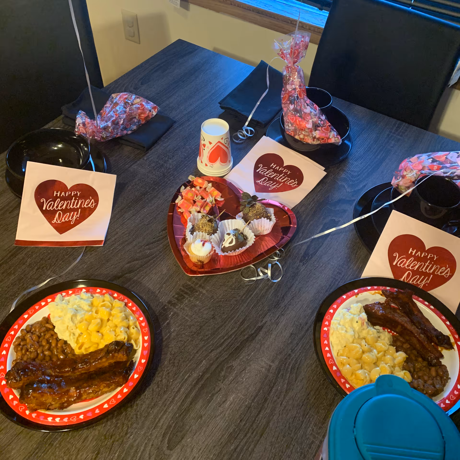 A dining table set for a Valentine's Day meal with two plates of food including ribs, baked beans, macaroni and cheese, and potato salad. The table is decorated with heart-themed napkins that say 'Happy Valentine's Day!', a heart-shaped tray with assorted desserts, and small gift bags wrapped in Valentine-themed cellophane. Black bowls and cups are also placed on the table.