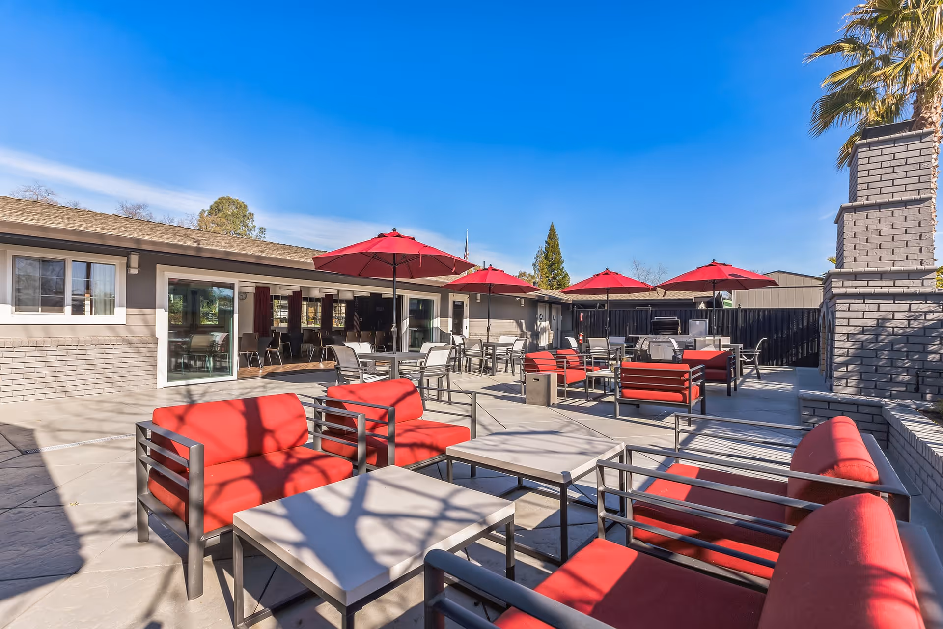 Outdoor patio area with multiple seating arrangements including red cushioned chairs and sofas, tables, and red umbrellas. The patio is adjacent to a building with large windows and sliding glass doors, under a clear blue sky.