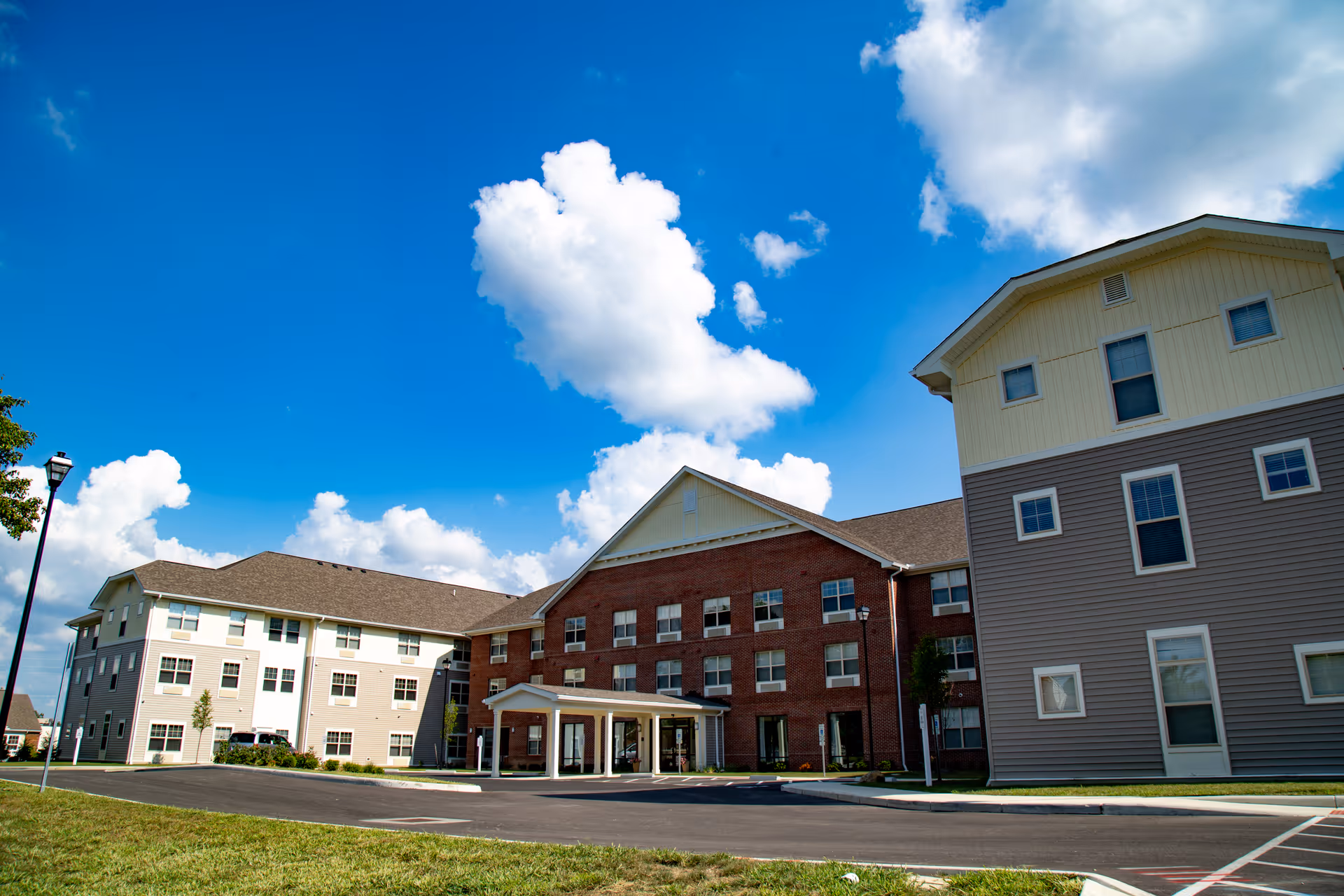 Exterior view of a multi-story senior living facility building under a bright blue sky with scattered white clouds. The building features a combination of brick and siding with multiple windows and a covered entrance.
