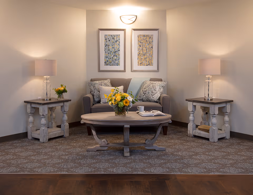 A cozy living room area with a gray sofa adorned with patterned and solid pillows, a wooden coffee table with a vase of yellow flowers, an open book, and a cup. Two matching wooden side tables with glass lamps and small flower arrangements flank the sofa. Two framed floral artworks hang on the wall above the sofa, illuminated by a wall sconce. The floor has a patterned carpet with a wooden border.